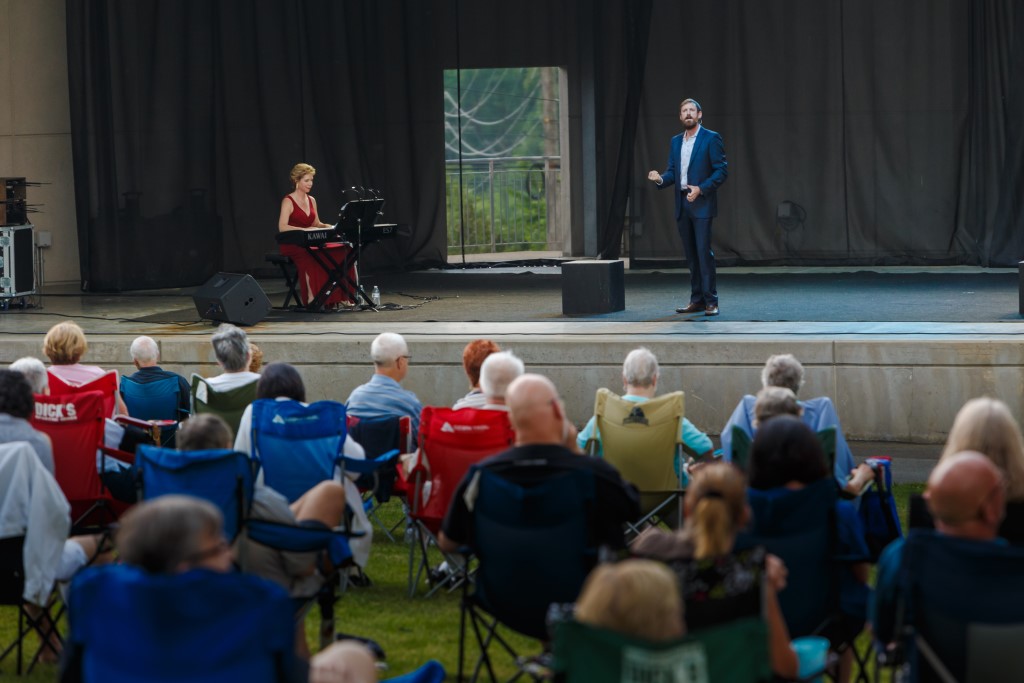 On stage a woman in a dress plays a keyboard while a man in a suit sings to an audience of people on camping chairs