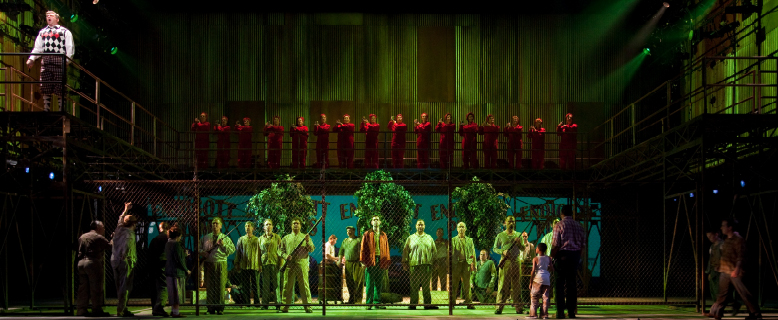 A group of men stand behind a chin-link fence with trees. Men with guns stand at the front of the fence. A group of people dressed in orange jumpsuits line a platform above the fenced-in area