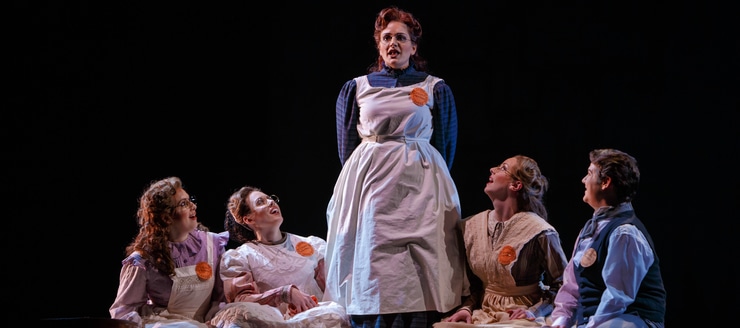 A woman in a dress stands up while three women in similar dresses sit and look up at her and a well-dressed man sits and looks up at her