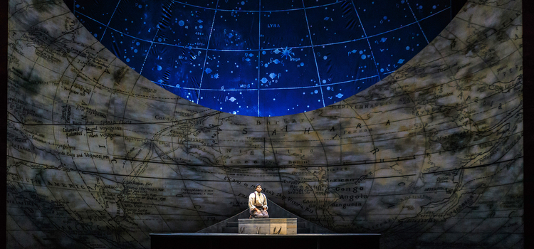 A man sits on his knees and stares out. The backdrop is a map with latitude and longitude lines displayed. 