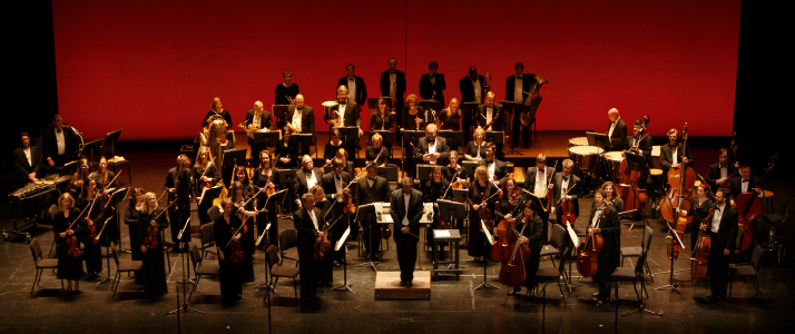 An entire orchestra posing for a picture in front of their chairs while holding their instruments 