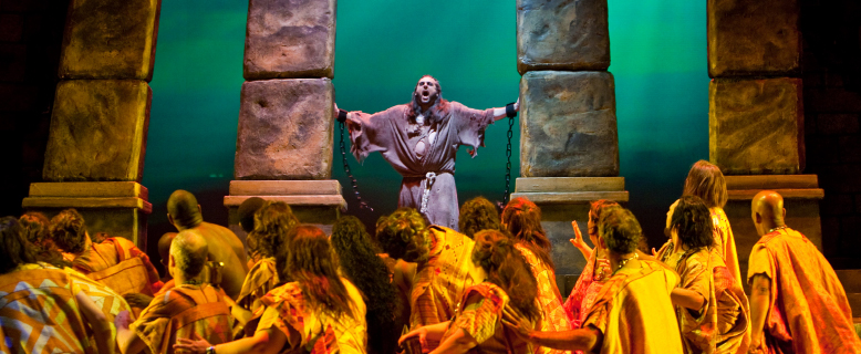 A man in scrappy clothes and chains stands between two stone pillars and sings while a group of people look up at him 