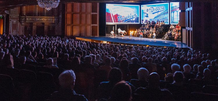 An audience in a large auditorium watching an opera with people in bleechers on the stage