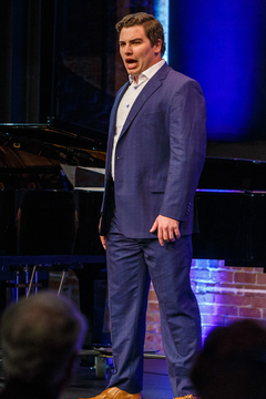 A well-dressed man sings in front of a crowd while standing in front of a camera