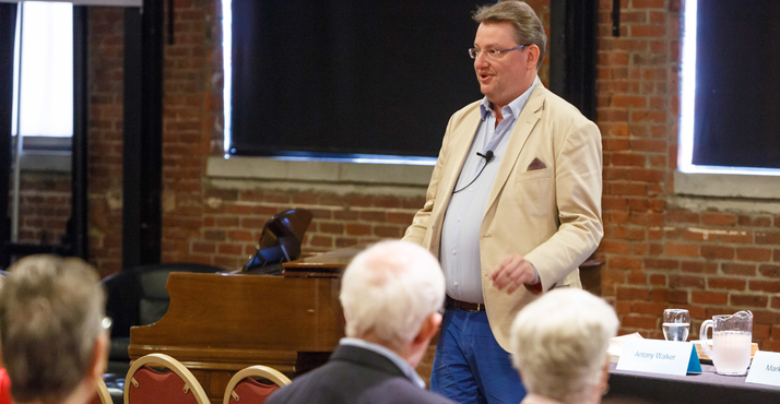 An old man talks in front of a group of people while standing 