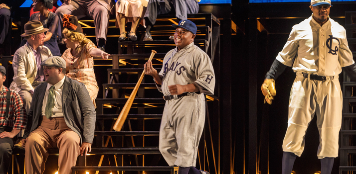 A man in a baseball uniform and hats twirls a bat in one hand while smiling. Another player stands to the side with a baseball mit on. Men and women in the bleachers behind them converse with each other