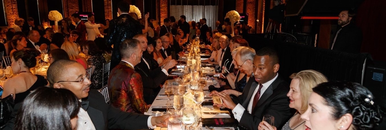 A long table with men and women in dresses and suits sitting on both sides and converse with each other