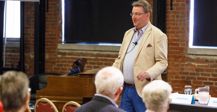 An old man talks in front of a group of people while standing